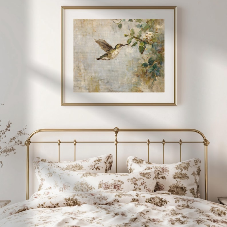 K&ouml;nnte beinhalten: Ein gerahmter Druck eines Kolibris im Flug, der sich einer bl&uuml;henden Zweig n&auml;hert. Das Kunstwerk weist weiche, ged&auml;mpfte Farben auf und wird &uuml;ber einem Bett mit floraler Bettw&auml;sche ausgestellt. Der Rahmen ist goldfarben.