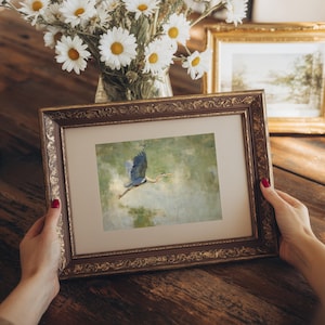 May include: A framed artwork featuring a blue heron in flight over water. The artwork is in a decorative, ornate brown and gold frame. A vase of white daisies and another framed picture are in the background.