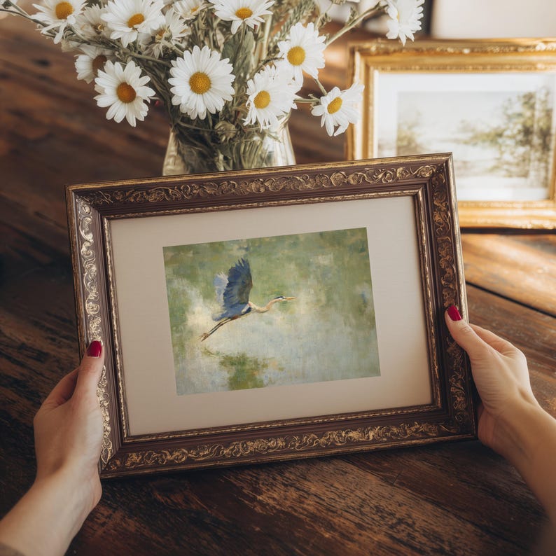 May include: A framed artwork featuring a blue heron in flight over water. The artwork is in a decorative, ornate brown and gold frame. A vase of white daisies and another framed picture are in the background.