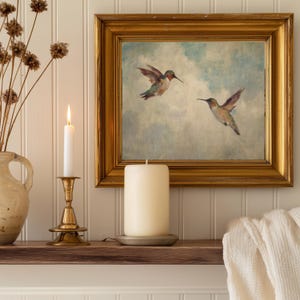May include: A gold-framed painting of two hummingbirds in flight hangs above a wooden shelf. A lit white candle sits in a brass candlestick, and a large pillar candle rests on a metal plate. Dried flowers are in a ceramic vase.