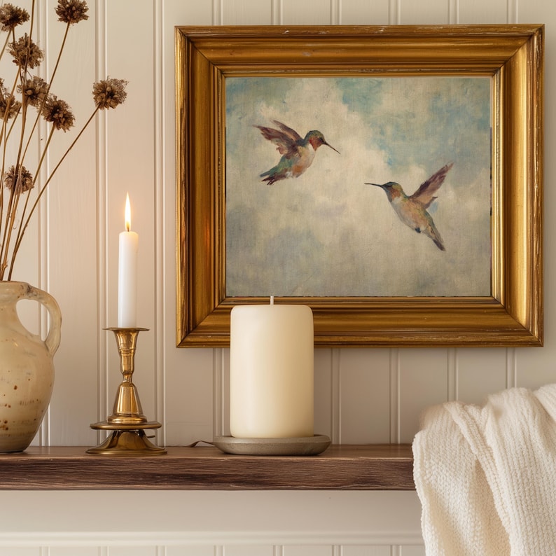 May include: A gold-framed painting of two hummingbirds in flight hangs above a wooden shelf. A lit white candle sits in a brass candlestick, and a large pillar candle rests on a metal plate. Dried flowers are in a ceramic vase.