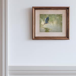 May include: A framed artwork featuring a painting of a blue heron in flight. The bird is depicted with outstretched wings against a soft, green and beige background. The frame is a warm, dark brown color, adding a classic touch to the artwork.