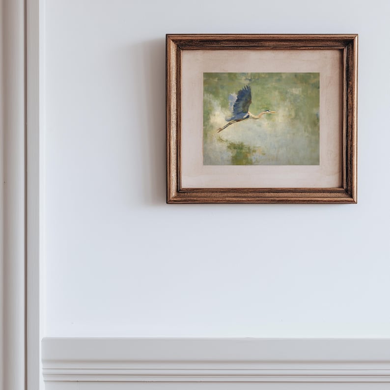 May include: A framed artwork featuring a painting of a blue heron in flight. The bird is depicted with outstretched wings against a soft, green and beige background. The frame is a warm, dark brown color, adding a classic touch to the artwork.