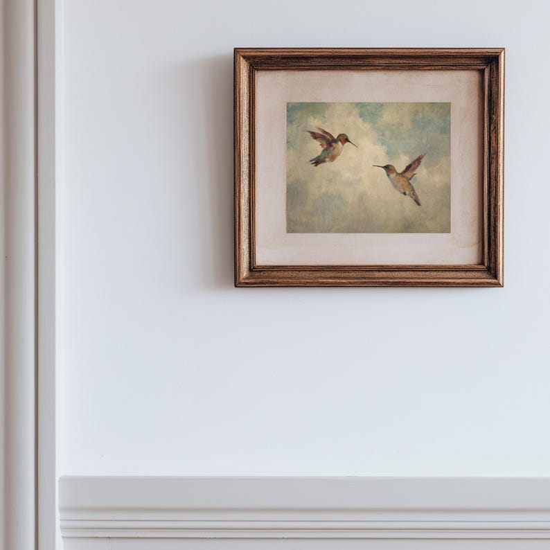 May include: A framed picture showing two hummingbirds in flight against a cloudy sky. The birds are depicted with brown and green feathers and long beaks. The frame is a dark, warm brown colour.