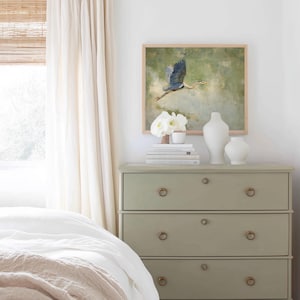 May include: A sage green dresser with three drawers, brass ring pulls, and a framed painting of a blue heron in flight. White vases and books are arranged on top, with a white curtain and a bed in the foreground.