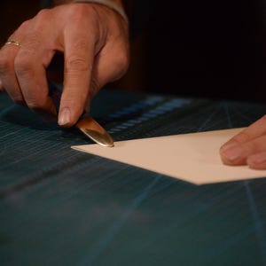Puede incluir: Un artesano utiliza una herramienta de borde dorada para recortar un trozo de papel de color claro. La herramienta se sujeta en ángulo y el papel descansa sobre una esterilla de corte verde oscuro. La escena está iluminada con luz cálida.