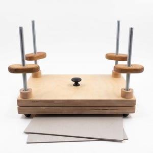 Könnte beinhalten: Eine Buchbinderpresse aus Holz mit zwei rechteckigen Sperrholzplatten, vier mit Gewinde versehenen Metallstangen mit verstellbaren Holzgriffen und einem zentralen schwarzen Knopf. Zwei rechteckige graue Kartonbögen liegen unter der Presse.