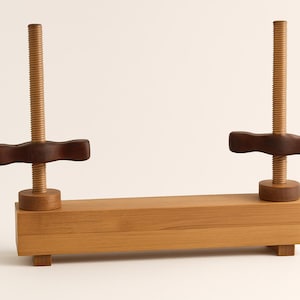 May include: A wooden book press featuring two vertical threaded rods with dark brown handles. The rectangular base has small feet. The wood has a light, natural finish, and the design is simple and functional.