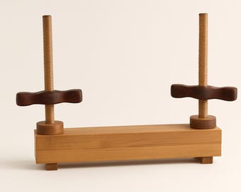 May include: A wooden book press with two vertical threaded rods and dark brown handles. The base is rectangular with small feet. The wood has a light, natural finish, and the design is simple and functional.