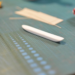 May include: A white bone folder tool rests on a green cutting mat, alongside a ruler and several thin, rectangular paper pieces. The bone folder is smooth and curved, used for paper crafting.