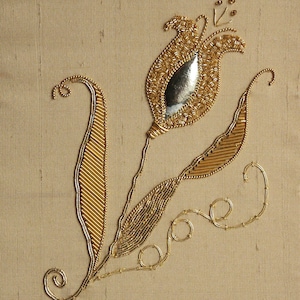 Könnte beinhalten: Goldene Perlenstickerei mit floralem Muster auf beigem Stoff. Das Design zeigt eine stilisierte Tulpe mit silbernem Zentrum, detaillierten Blättern und zarten Ranken. Die aufwendige Perlenarbeit erzeugt einen strukturierten, dreidimensionalen Effekt.