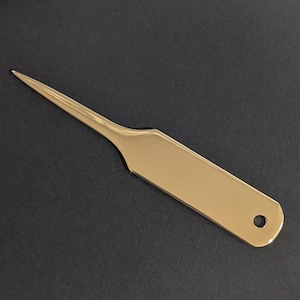 May include: A gold-coloured letter opener with a long, pointed blade and a rectangular handle. The handle has a small hole near the end. The letter opener is made of a shiny, reflective material.