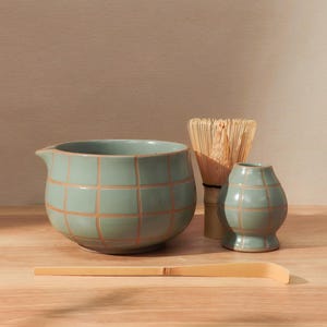 May include: A matcha tea set featuring a celadon green bowl with a spout, a small vase, and a bamboo whisk. The bowl and vase have a grid pattern. A bamboo scoop rests in front of the set on a wooden surface.