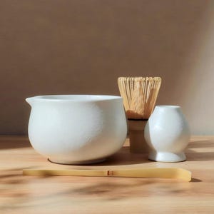 May include: A white ceramic matcha bowl with a spout, a bamboo whisk, a small white vase, and a bamboo scoop. The items are arranged on a wooden surface, suggesting a tea ceremony setup.