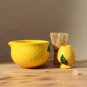 May include: A yellow ceramic matcha bowl, whisk holder, and bamboo whisk set. The bowl has a textured surface and a spout. The whisk holder is shaped like a lemon. A bamboo scoop rests in front of the bowl.