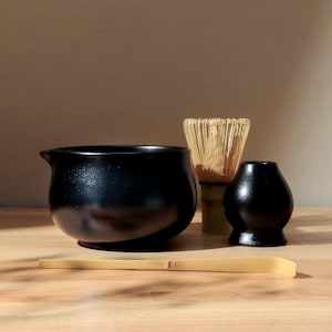 Puede incluir: Un juego de té matcha negro que incluye un cuenco con pico, un batidor de bambú, un recipiente para té negro y una cuchara de bambú. El juego está dispuesto sobre una superficie de madera clara.