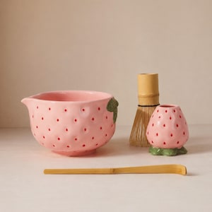 May include: A pink ceramic matcha bowl, strawberry-shaped holder, and bamboo whisk set. The bowl and holder feature red dot details. A bamboo scoop rests in front of the set. The set is designed for preparing matcha tea.