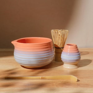 Puede incluir: Un juego de té matcha de cerámica con un cuenco degradado en melocotón y azul, un batidor de bambú, un pequeño soporte para batidor de cerámica y una cuchara de bambú. El juego está dispuesto sobre una superficie de madera.