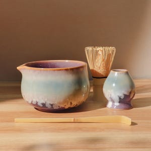 May include: A matcha tea set comprising a ceramic bowl with a spout, a bamboo whisk, a small ceramic holder, and a bamboo scoop. The bowl has a gradient of purple, blue, and brown hues. The whisk is made of thin bamboo strips.