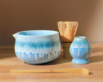 Puede incluir: Un juego de té matcha con un cuenco de cerámica azul claro con pico, un batidor de bambú, un pequeño recipiente de cerámica azul y una cuchara de bambú. El cuenco y el recipiente tienen un diseño de esmalte goteado blanco y azul.