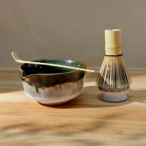 Op de afbeelding: Een matcha-theeset met een keramische kom met een groene en bruine glazuur, een bamboe klopper en een bamboe lepel. De kom staat op een houten oppervlak, met de klopper en lepel in de buurt. De set is ontworpen voor het bereiden van matcha thee.
