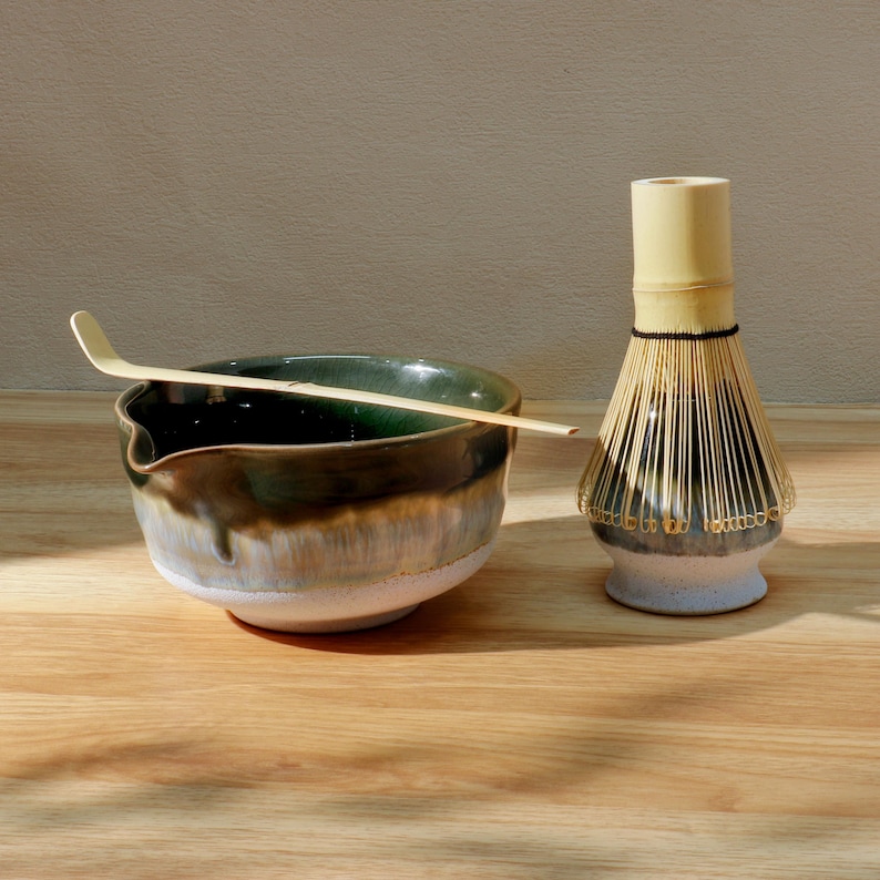 Op de afbeelding: Een matcha-theeset met een keramische kom met een groene en bruine glazuur, een bamboe klopper en een bamboe lepel. De kom staat op een houten oppervlak, met de klopper en lepel in de buurt. De set is ontworpen voor het bereiden van matcha thee.