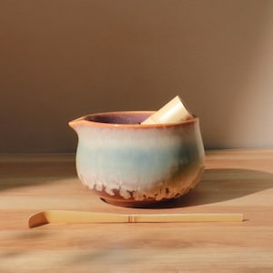 May include: A ceramic matcha bowl with a spout, featuring a gradient of colors from brown to blue and purple. A bamboo whisk and a bamboo scoop rest beside the bowl. The bowl has a unique, textured glaze.