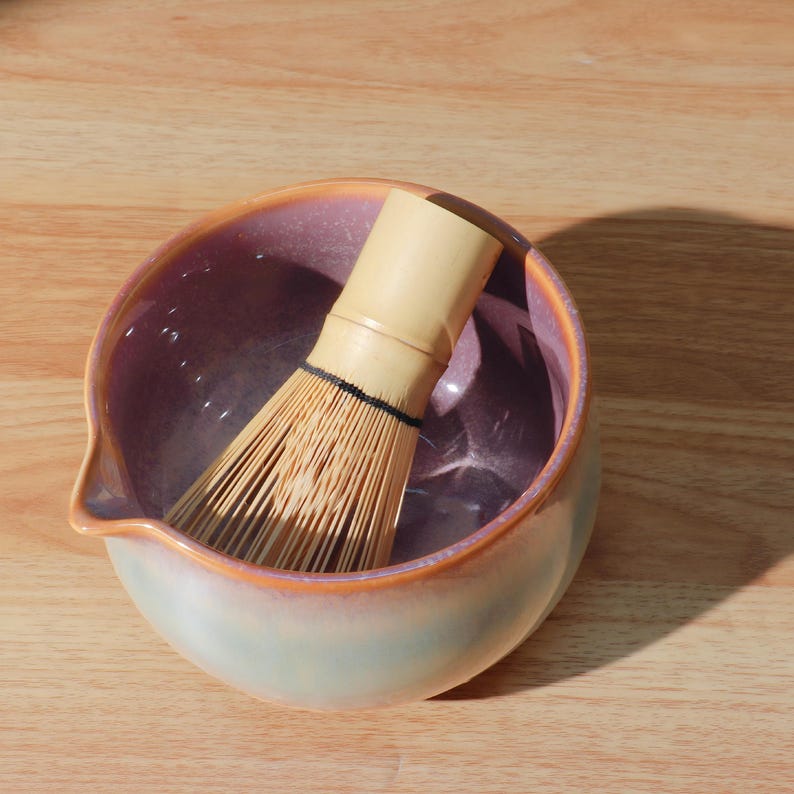 May include: A ceramic matcha bowl with a light purple interior and a light green and orange exterior. A bamboo matcha whisk rests inside the bowl. The bowl has a small spout on the side. The whisk is made of thin bamboo tines.