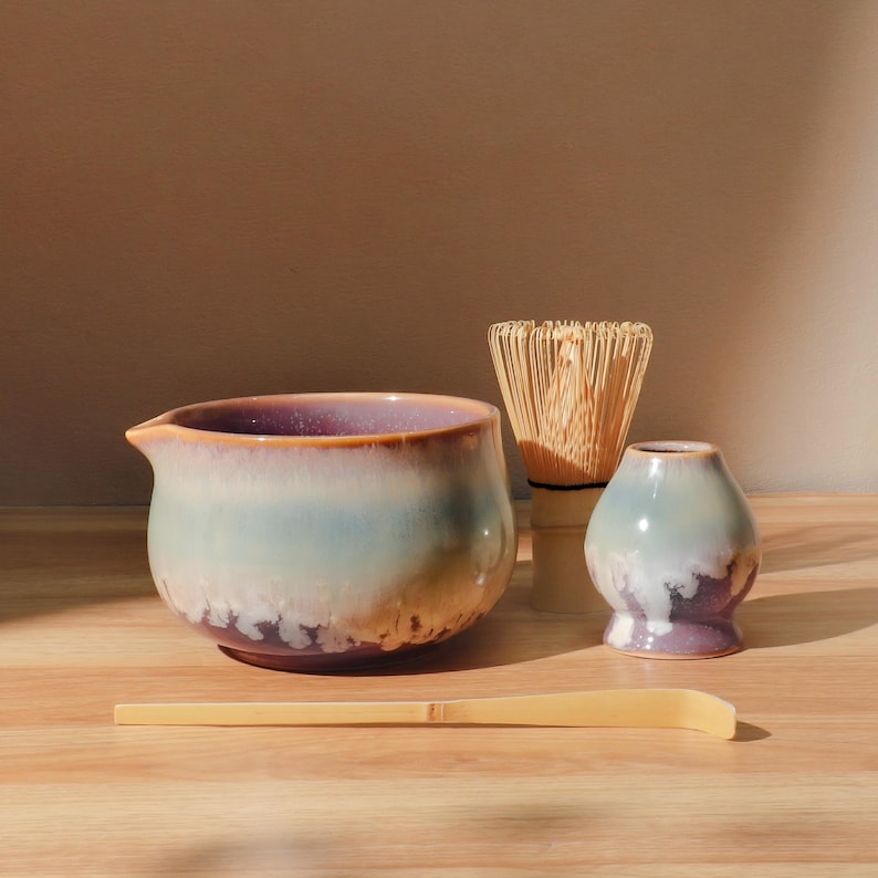 May include: A matcha tea set featuring a ceramic bowl with a spout, a bamboo whisk, a small ceramic holder, and a bamboo scoop. The bowl has a gradient of colors, including purple, blue, and brown. The whisk is made of thin bamboo strips.