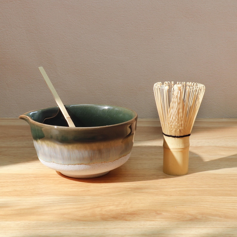 Op de afbeelding: Een matcha-theeset met een keramische kom met een tuit, een bamboe klopper en een bamboe lepel. De kom heeft een verloop van groen, bruin en wit. De klopper is gemaakt van dunne bamboeharen.