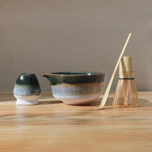 Op de afbeelding: Een matcha-theeset met een keramische kom, een kleine beker, een bamboe klopper en een bamboe lepel. De keramische stukken hebben een kleurengradi&euml;nt van wit, blauw en groen. De klopper en lepel zijn gemaakt van lichtgekleurd bamboe.