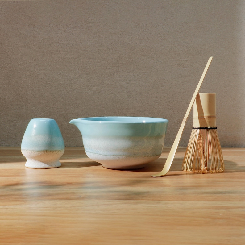 Op de afbeelding: Een matcha-theeset met een lichtblauwe keramische kom, een garde, een lepel en een kleine keramische container. De kom heeft een tuit en een witte basis. De garde is van bamboe. De set staat op een houten oppervlak.