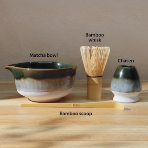 Op de afbeelding: Een matcha-theeset met een matcha-kom van keramiek, een bamboe klopper, een chasen en een bamboelepel. De kom heeft een donkergroen en wit glazuur. De bamboe klopper en lepel zijn lichtbruin.