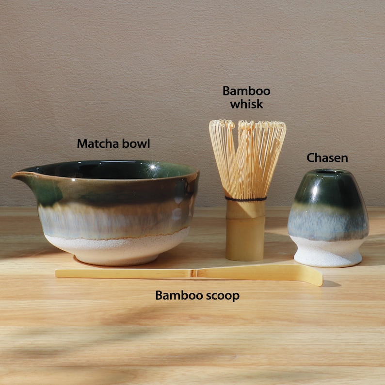 Op de afbeelding: Een matcha-theeset met een matcha-kom van keramiek, een bamboe klopper, een chasen en een bamboelepel. De kom heeft een donkergroen en wit glazuur. De bamboe klopper en lepel zijn lichtbruin.