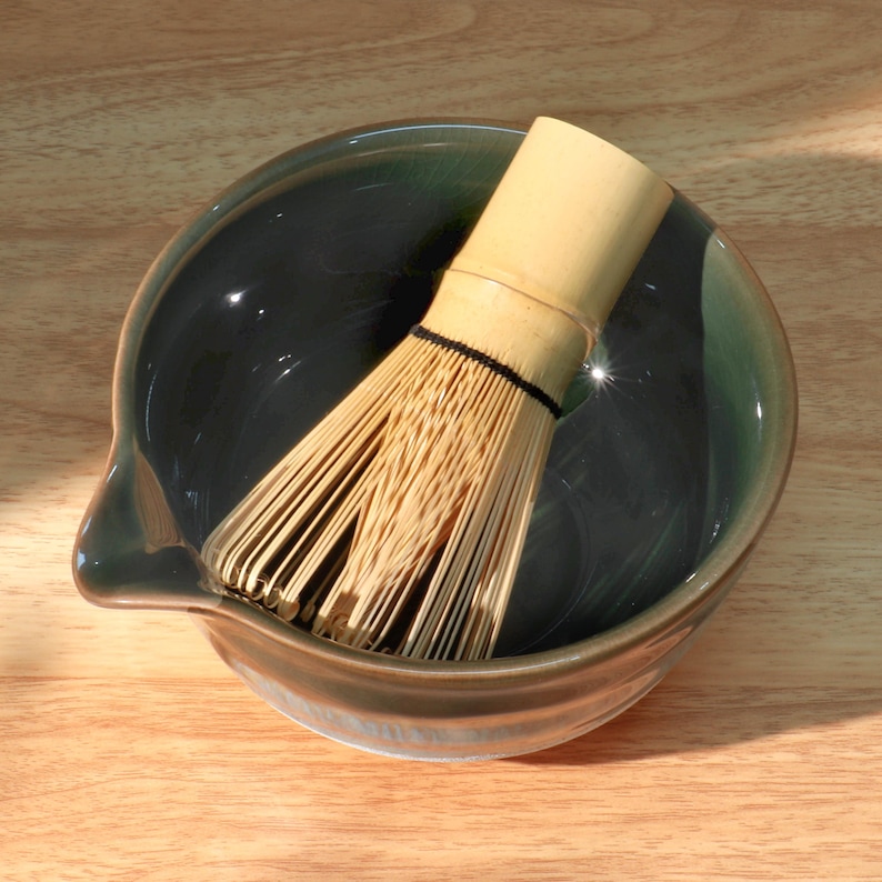 Op de afbeelding: Een matcha-theeset met een donkergroene keramische kom met een tuit en een bamboe klopper. De klopper, gemaakt van dun, lichtgekleurd bamboe, bevindt zich in de kom, klaar voor gebruik.