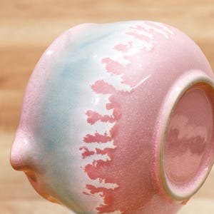 May include: A ceramic bowl with a unique design. The bowl features a gradient of colors, including pink, blue, and white. The pink color dominates the right side, transitioning to blue and white on the left. The bowl has a textured surface and a small spout.