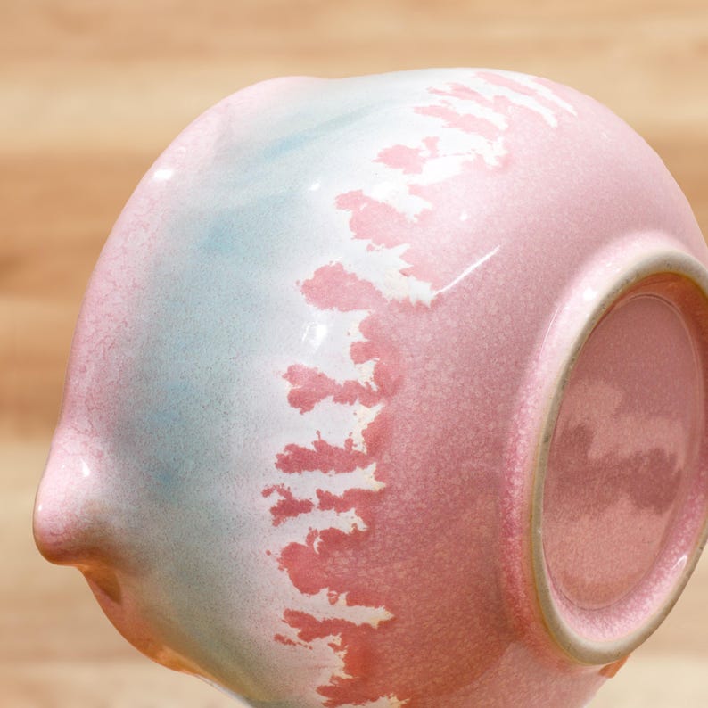 May include: A ceramic bowl with a unique design. The bowl features a gradient of colors, including pink, blue, and white. The pink color dominates the right side, transitioning to blue and white on the left. The bowl has a textured surface and a small spout.