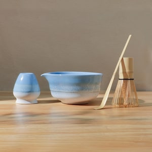 Op de afbeelding: Een matcha-theeset met een lichtblauwe keramische kom, een garde, een lepel en een kleine blauw-witte keramische houder. De kom heeft een tuit. De garde is gemaakt van bamboe. De set staat op een houten oppervlak.