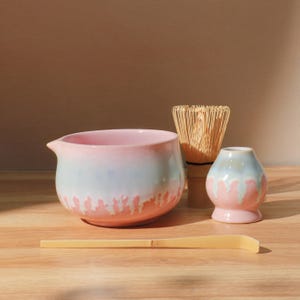 May include: A matcha tea set featuring a pink and blue ceramic bowl, a bamboo whisk, a small vase, and a bamboo scoop. The bowl has a spout and a drip-like design. The set is arranged on a wooden surface.