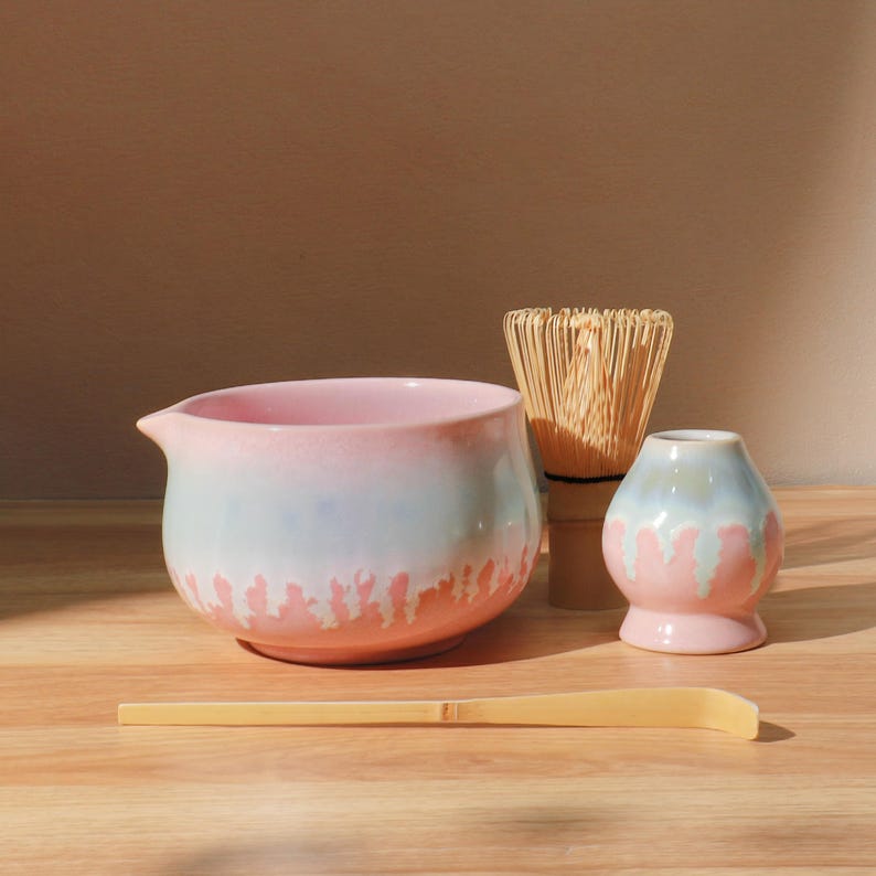 May include: A matcha tea set featuring a pink and blue ceramic bowl, a bamboo whisk, a small vase, and a bamboo scoop. The bowl has a spout and a drip-like design. The set is arranged on a wooden surface.