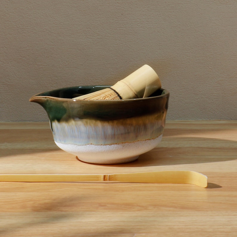 Op de afbeelding: Een keramische matcha kom met een groene en bruine glazuur, een bamboe klopper en een bamboe lepel. De kom heeft een tuit om gemakkelijk te schenken. De klopper zit in de kom. De lepel ligt op het houten oppervlak.