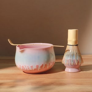 May include: A matcha tea set featuring a pink and blue ceramic bowl with a wooden scoop and a bamboo whisk. The bowl has a unique, textured design along the bottom edge. The whisk has a light yellow handle and a bundle of bamboo bristles.