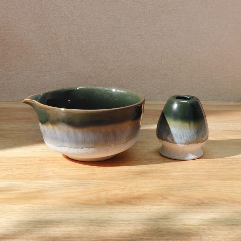 Op de afbeelding: Een keramische matcha kom en garde houder set. De kom is diepgroen met een tuit en een kleurovergang, van groen naar wit. De garde houder heeft een vergelijkbare kleurovergang, met een afgeronde vorm.