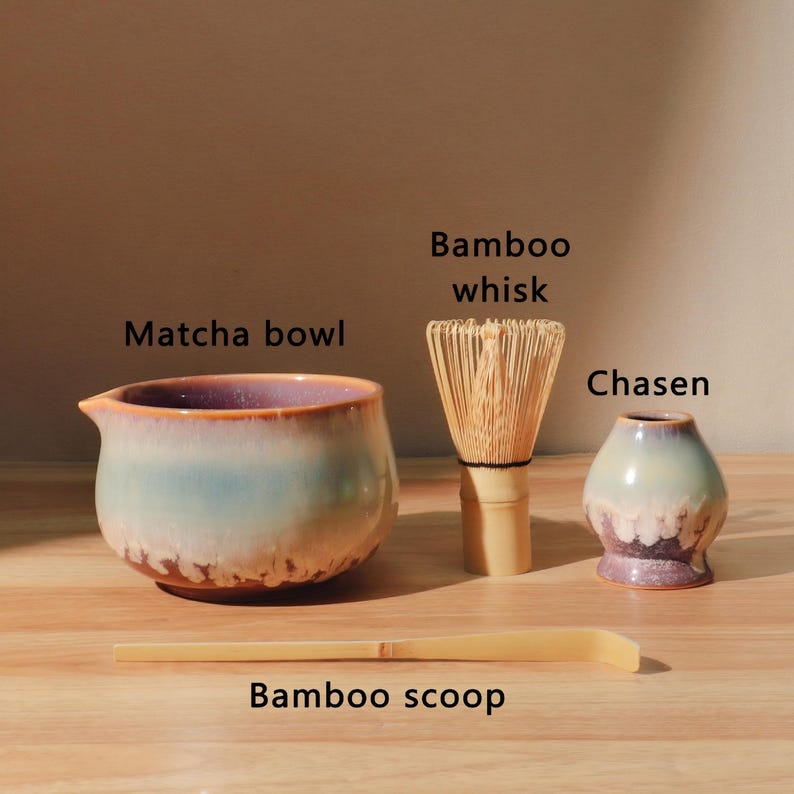 May include: A matcha tea set with a blue and brown ceramic matcha bowl, a bamboo whisk, a chasen, and a bamboo scoop. The text labels the items as "Matcha bowl", "Bamboo whisk", "Chasen", and "Bamboo scoop".