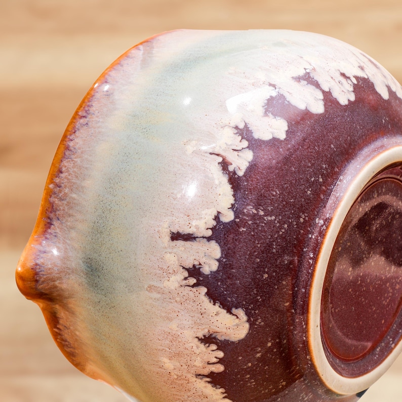 May include: A ceramic bowl with a unique glaze. The bowl features a blend of colors, including a deep burgundy, creamy white, and a touch of light green. The glaze has a flowing, organic pattern, creating a visually appealing texture. The rim is a warm orange.