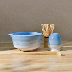 May include: A light blue ceramic matcha bowl with a spout, a bamboo whisk, a blue and white ceramic matcha tea holder, and a bamboo scoop. The items are arranged on a wooden surface.