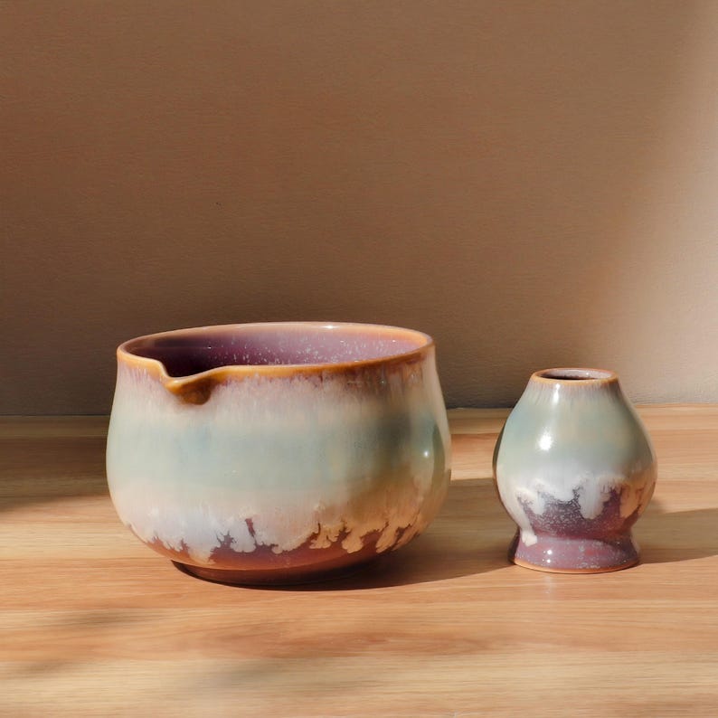 May include: A ceramic tea bowl and whisk holder set. The bowl is wide with a spout, featuring a gradient of colors from purple to light blue and brown. The whisk holder is small, with a similar color gradient. Both are on a wooden surface.