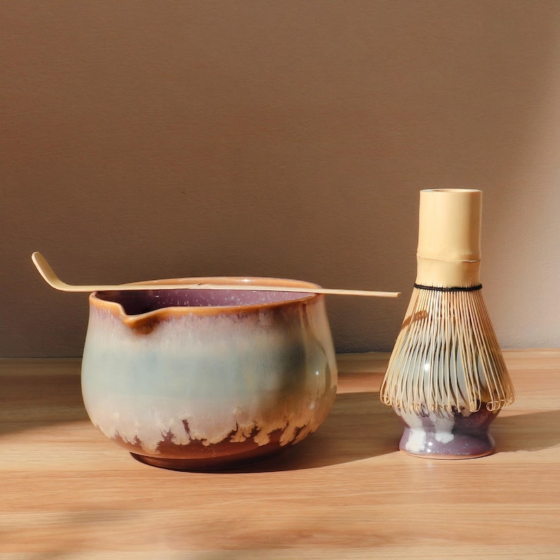 May include: A matcha tea set featuring a ceramic bowl with a light blue and brown glaze, a bamboo whisk, and a bamboo scoop. The bowl has a purple interior and sits on a wooden surface. The whisk has a light yellow handle and a purple base.