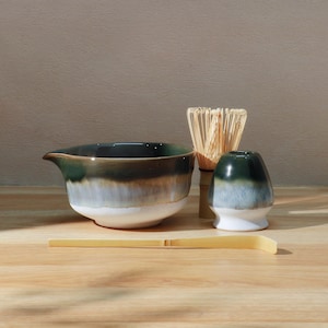 Könnte beinhalten: Ein Matcha-Teeset mit einer Keramikschale, einem Schneebesen, einem Schneebesenhalter und einem Bambuslöffel. Die Schale hat eine dunkelgrüne, braune und weiße Glasur. Der Schneebesen ist aus Bambus, und der Schneebesenhalter ist dunkelgrün und weiß.