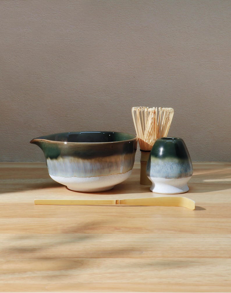 Op de afbeelding: Een matcha-theeset met een keramische kom, een klopper, een klopperhouder en een bamboelepel. De kom heeft een donkergroen, bruin en wit glazuur. De klopper is gemaakt van bamboe en de klopperhouder is donkergroen en wit.